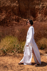Woman in a white dress standing in a desert-like landscape with a rocky wall in the background.