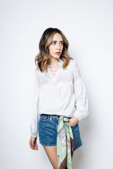 A woman modeling a white blouse with long blouson sleeves, a V-shaped keyhole neckline with ties, and a high pleated collar. She is also wearing blue denim shorts and a scarf tied around the waist.