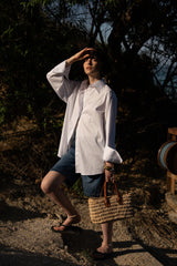 Person in a white shirt and denim shorts standing outdoors with a straw bag.