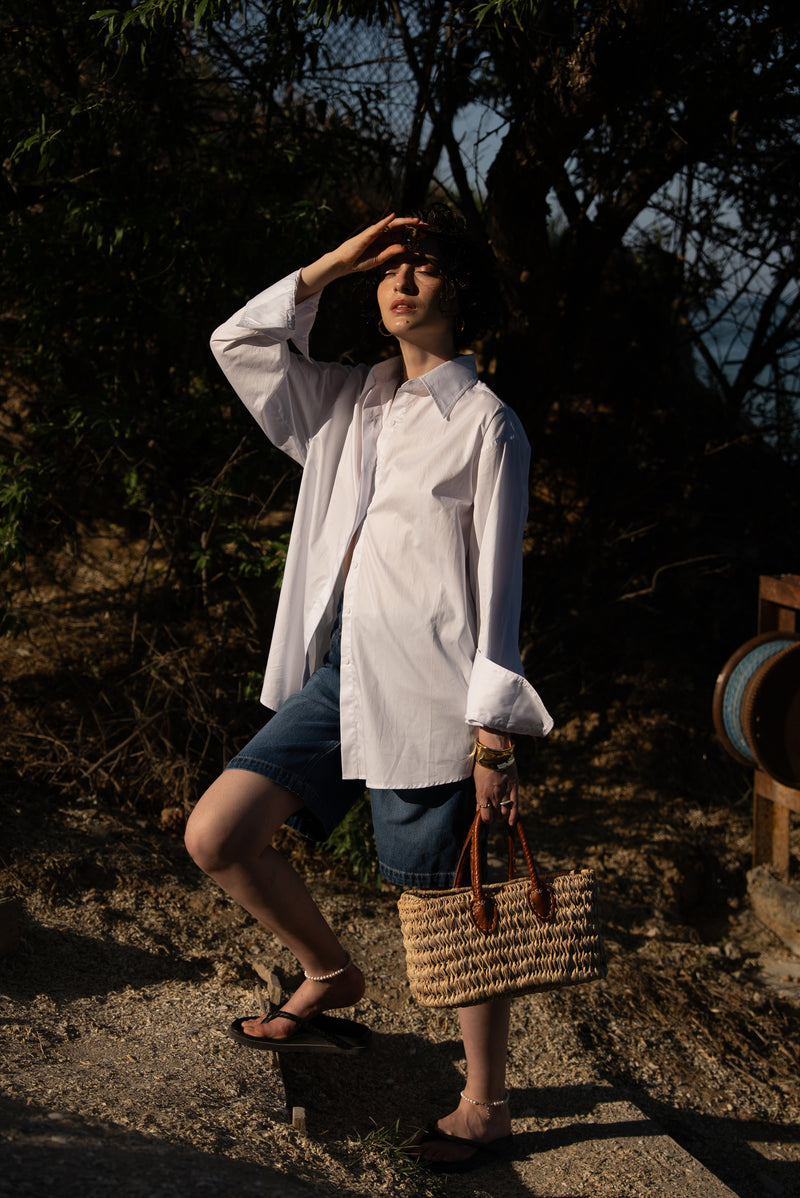 Person in a white shirt and denim shorts standing outdoors with a straw bag.