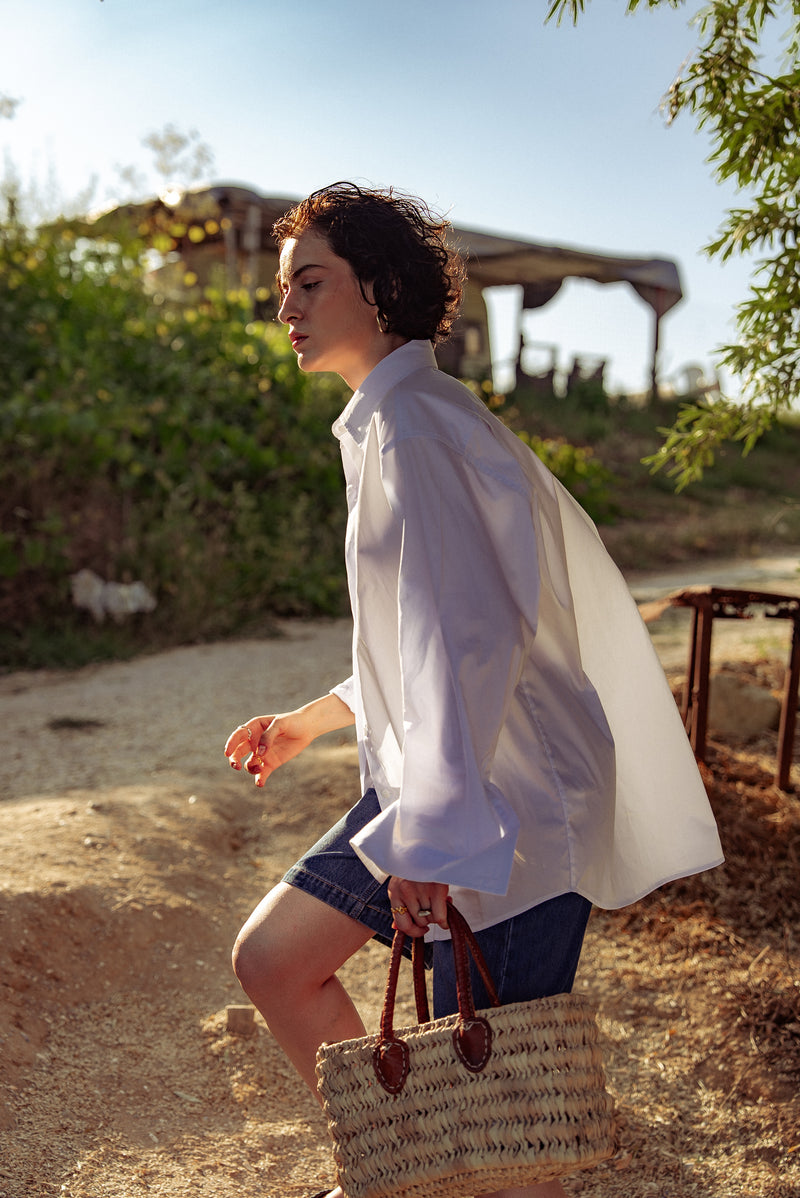 Woman in a white shirt and denim shorts holding a straw bag outdoors.