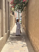 A woman wearing a long white abaya and sunglasses stands in a narrow sunlit alleyway between beige stucco walls, with a flowering tree overhead.