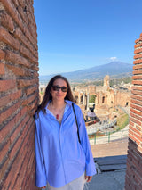 A woman wearing a light blue button-up shirt and sunglasses stands in a narrow brick corridor with a scenic view of ancient ruins and Mount Vesuvius in the background under a clear blue sky.