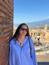 A woman wearing a light blue button-up shirt and sunglasses stands outdoors against a brick wall with ancient ruins and a mountain in the background under a clear blue sky.